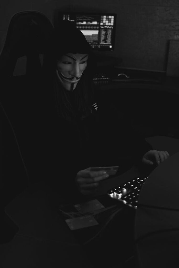 A hacker wearing a Guy Fawkes mask using a credit card in a dark room, symbolizing cybercrime.